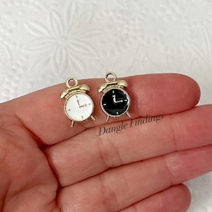 May include: Two gold-tone alarm clock charms, one with a white face and one with a black face. Both charms have black hands and a gold-tone ring around the face.