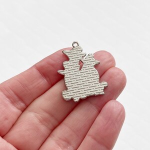 Pode incluir: Encanto de prata com a silhueta de dois gatos sentados de costas um para o outro. O encanto tem uma superf&iacute;cie texturizada.