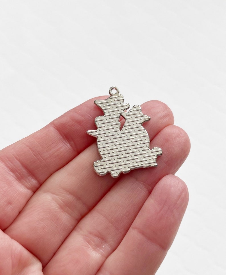 Pode incluir: Encanto de prata com a silhueta de dois gatos sentados de costas um para o outro. O encanto tem uma superf&iacute;cie texturizada.