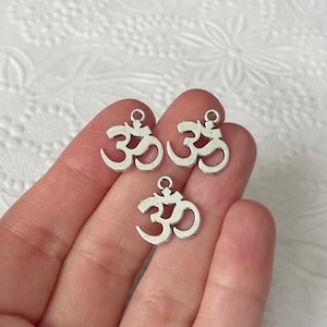 May include: Three silver-colored Om symbol charms. Each charm features the Om symbol with a small loop at the top for attaching to jewelry or other crafts. The charms are displayed against a white background.