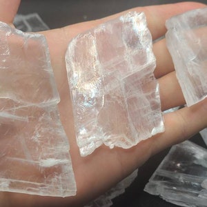 May include: A collection of clear selenite crystal pieces with a layered, translucent appearance. The crystals are various sizes and shapes, with some having a rectangular shape.