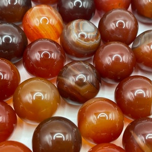 May include: A collection of carnelian gemstone spheres in various shades of red, orange, and brown. The spheres are smooth and polished, with natural banding and patterns.