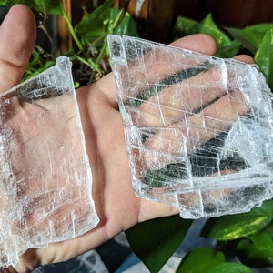 May include: Two pieces of clear selenite crystal, one larger than the other, with a layered, striated texture. The crystals are held in a hand.