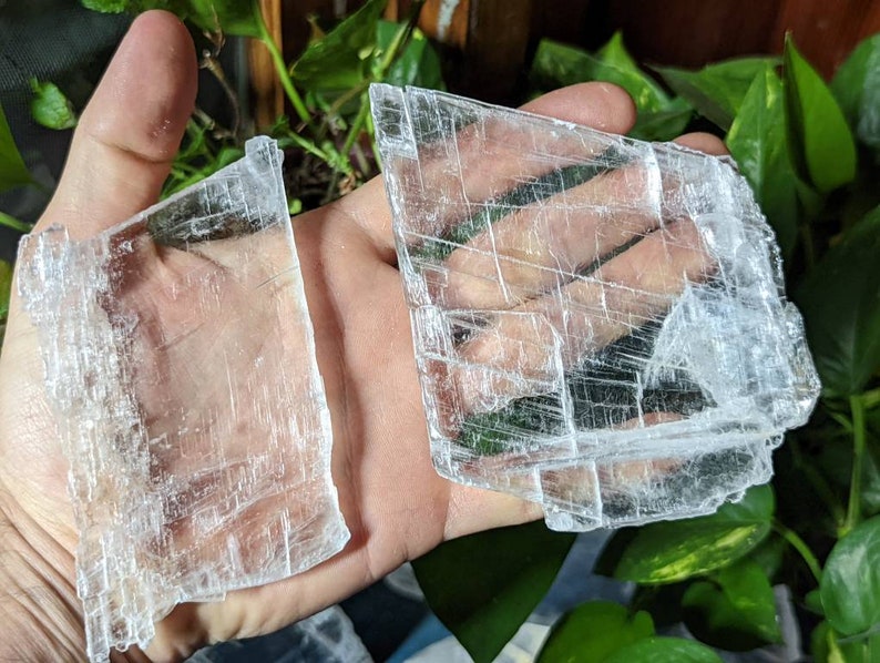 May include: Two pieces of clear selenite crystal, one larger than the other, with a layered, striated texture. The crystals are held in a hand.