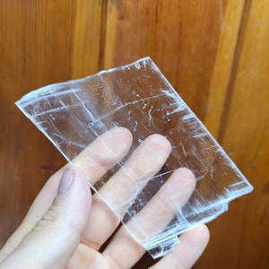 May include: A clear, translucent selenite crystal slab with visible striations. The crystal is held in a hand, showcasing its size and texture.
