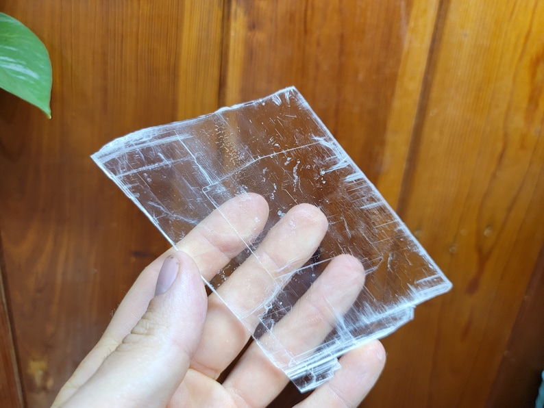 May include: A clear, translucent selenite crystal slab with visible striations. The crystal is held in a hand, showcasing its size and texture.