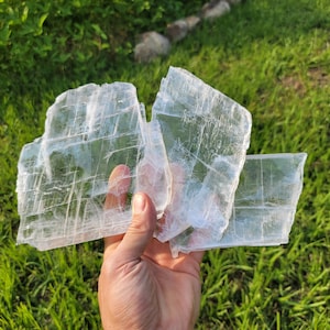 May include: Three pieces of clear selenite crystal, stacked on top of each other, with a hand holding the bottom piece. The crystals are translucent and have a layered, striated appearance.