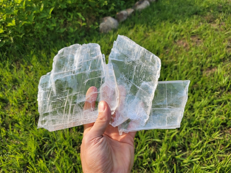 May include: Three pieces of clear selenite crystal, stacked on top of each other, with a hand holding the bottom piece. The crystals are translucent and have a layered, striated appearance.