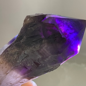 Könnte beinhalten: Ein großer, violetter Amethystkristall mit einer spitzen Spitze. Der Kristall hat oben eine dunkelviolette Farbe und verblasst nach unten zu einem helleren Violett. Der Kristall hat eine raue, natürliche Textur.