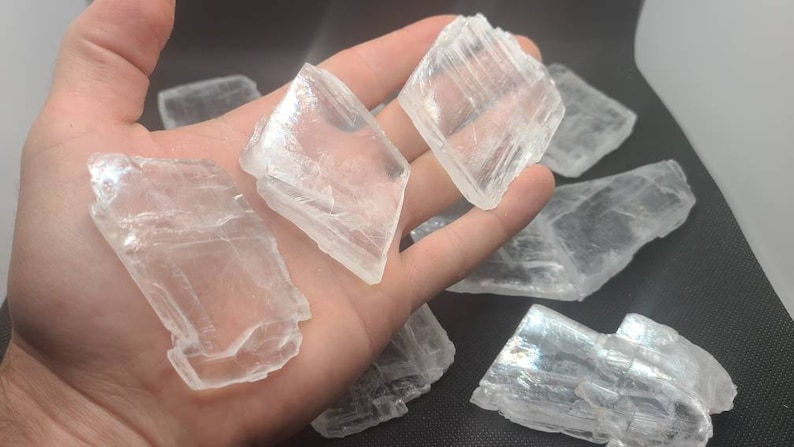 May include: A collection of selenite crystals, also known as satin spar, with a translucent, white appearance. The crystals have a layered, striated texture and are arranged on a dark surface.