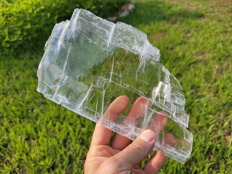 May include: A large, clear selenite crystal with a layered, striated texture. The crystal is translucent and has a slight sheen. It is held in a hand against a background of green grass.