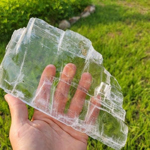 May include: A large, clear selenite crystal slab with a layered, translucent appearance. The crystal is held in a hand, showcasing its size and delicate structure.