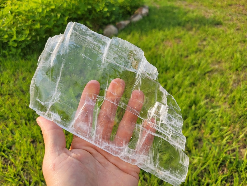 May include: A large, clear selenite crystal slab with a layered, translucent appearance. The crystal is held in a hand, showcasing its size and delicate structure.