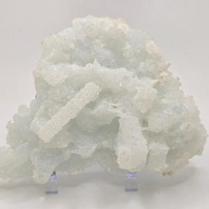 Może przedstawiać: Jasnoniebieski okaz minerału o krystalicznej strukturze. Minerał ma szorstką, teksturowaną powierzchnię i jest zamontowany na przezroczystym stojaku. Ogólny kształt jest nieregularny i organiczny, z skupiskiem formacji kryształowych.
