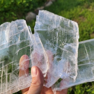 May include: Three large, clear, rectangular selenite crystals with visible striations. The crystals are stacked on top of each other and are being held in a hand.