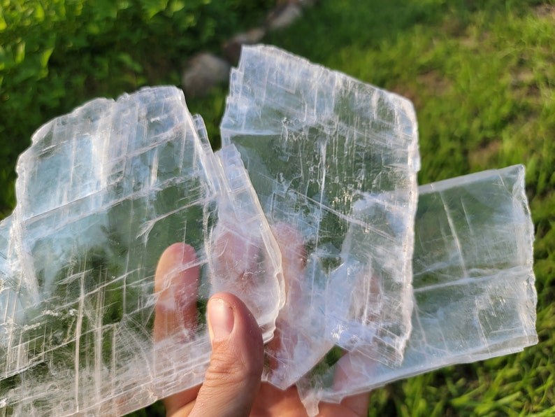 May include: Three large, clear, rectangular selenite crystals with visible striations. The crystals are stacked on top of each other and are being held in a hand.