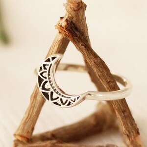 May include: A silver ring with a crescent moon design. The moon is decorated with a detailed mandala pattern. The ring is displayed on a wooden stand.