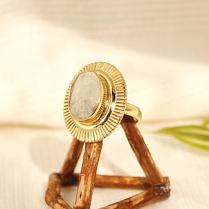 May include: A gold ring with a white stone set in a sunburst design. The ring is displayed on a wooden stand.