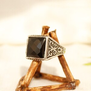 May include: A silver ring with a black square gemstone and intricate filigree design. The ring is displayed on a wooden stand.