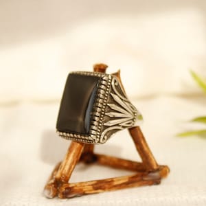 May include: A silver ring with a black square stone set in a decorative silver band. The ring is displayed on a wooden stand.