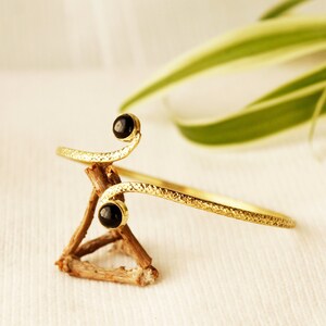 May include: A gold-toned bracelet with a textured finish and two black gemstone accents. The bracelet is curved and has a delicate design.