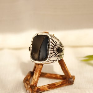 May include: A silver ring with a black stone set in a square shape. The ring has a detailed design around the stone, resembling a flower.