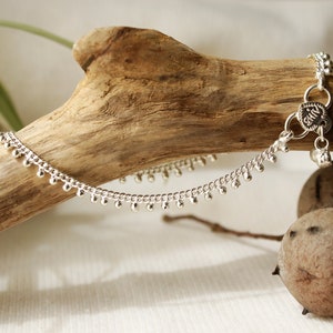 Può includere: Cavigliera d'argento con un ciondolo a forma di cuore e una catena pendente di piccole perle.