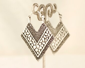 May include: A pair of silver hoop earrings with a geometric, diamond-shaped design. The earrings have a textured surface with a pattern of small, raised circles.