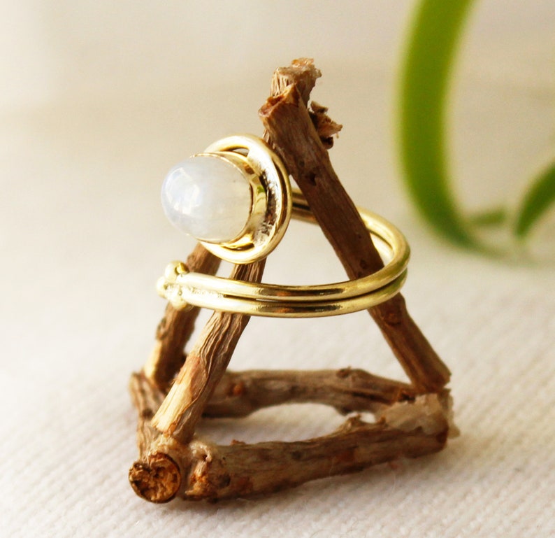 K&ouml;nnte beinhalten: Ein goldener Ring mit einem wei&szlig;en Mondstein, der in einer Zargenfassung gefasst ist. Der Ring befindet sich auf einem h&ouml;lzernen Ringst&auml;nder.