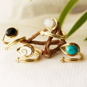 K&ouml;nnte beinhalten: Vier goldene Ringe mit verschiedenen farbigen Steinen. Die Ringe sind um ein St&uuml;ck Treibholz gewickelt. Die Steine sind schwarz, wei&szlig;, t&uuml;rkis und eine wei&szlig;e Muschel mit einer braunen Spirale.