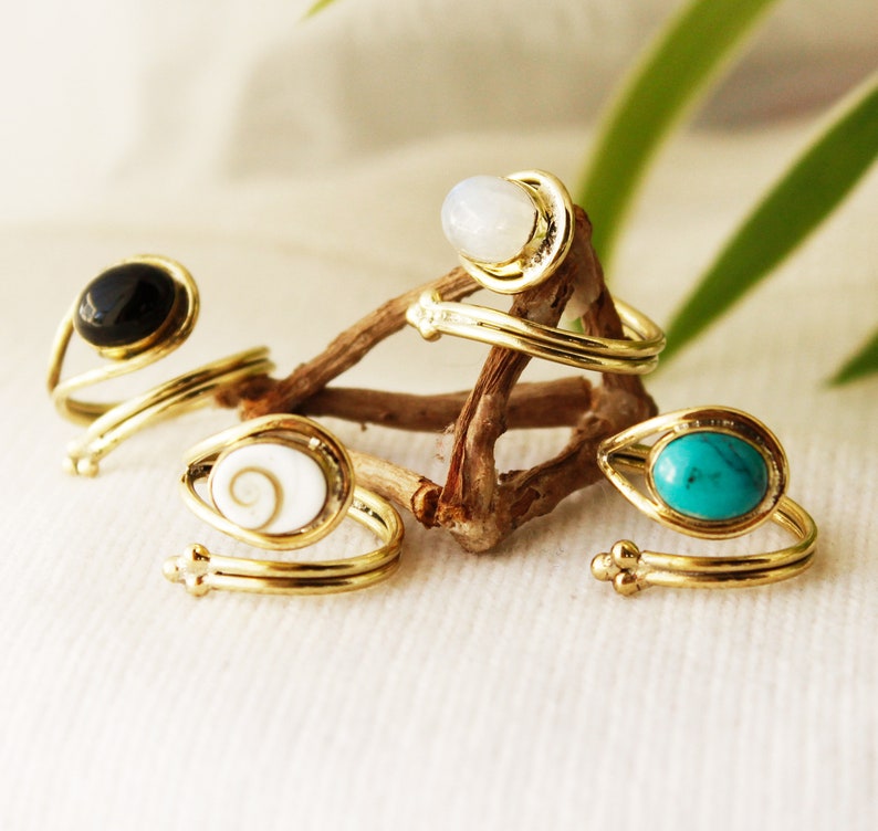K&ouml;nnte beinhalten: Vier goldene Ringe mit verschiedenen farbigen Steinen. Die Ringe sind um ein St&uuml;ck Treibholz gewickelt. Die Steine sind schwarz, wei&szlig;, t&uuml;rkis und eine wei&szlig;e Muschel mit einer braunen Spirale.