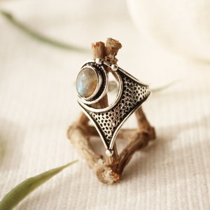 May include: A silver ring with a large, oval-shaped moonstone set in a geometric design. The ring is on a wooden stand.