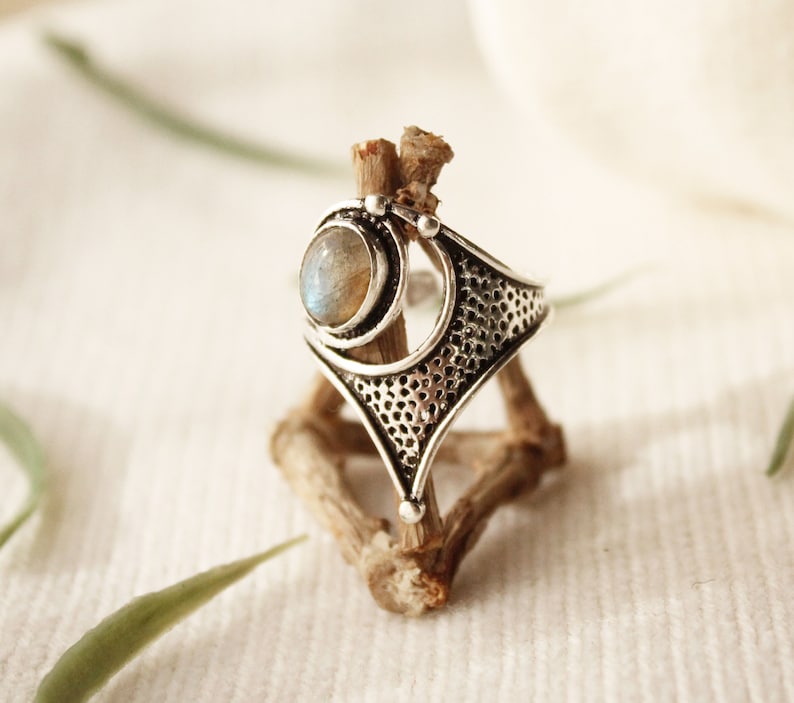 May include: A silver ring with a large, oval-shaped moonstone set in a geometric design. The ring is on a wooden stand.
