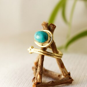K&ouml;nnte beinhalten: Ein goldener Ring mit einem t&uuml;rkisfarbenen Stein, der in einem gedrehten Band gefasst ist. Der Ring wird auf einem Holzst&auml;nder pr&auml;sentiert.