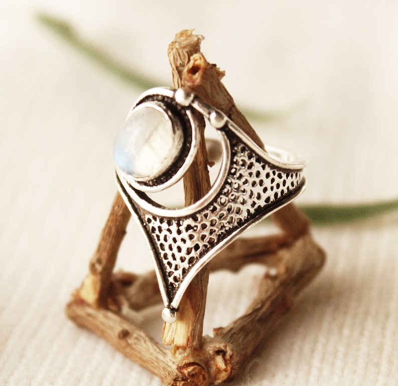 May include: A silver ring with a large, round, white moonstone set in a geometric design. The ring is displayed on a wooden stand.