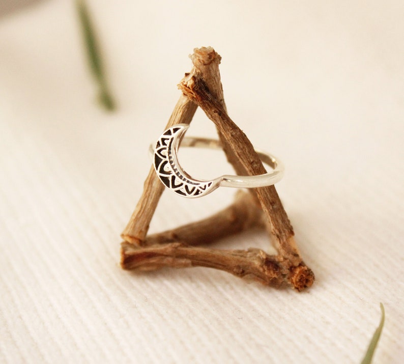 May include: A silver ring with a crescent moon design. The moon is decorated with a detailed mandala pattern. The ring is displayed on a wooden stand.