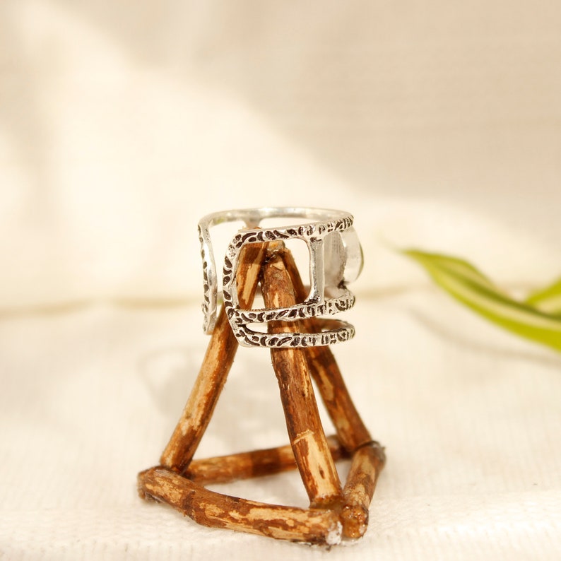 May include: Silver ring with an intricate design and a clear stone. The ring is displayed on a wooden stand.