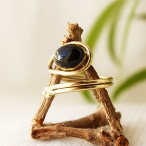 K&ouml;nnte beinhalten: Ein goldener Ring mit einem schwarzen ovalen Edelstein, der in einem drahtgewickelten Design gefasst ist. Der Ring wird auf einem Holzst&auml;nder pr&auml;sentiert.