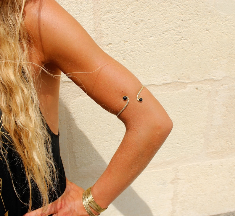 May include: A gold arm cuff with two black beads. The cuff is worn on the upper arm.