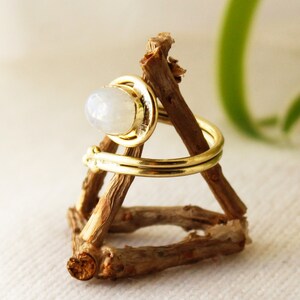 K&ouml;nnte beinhalten: Ein goldener Ring mit einem wei&szlig;en Mondstein, der in einer Zargenfassung gefasst ist. Der Ring befindet sich auf einem h&ouml;lzernen Ringst&auml;nder.