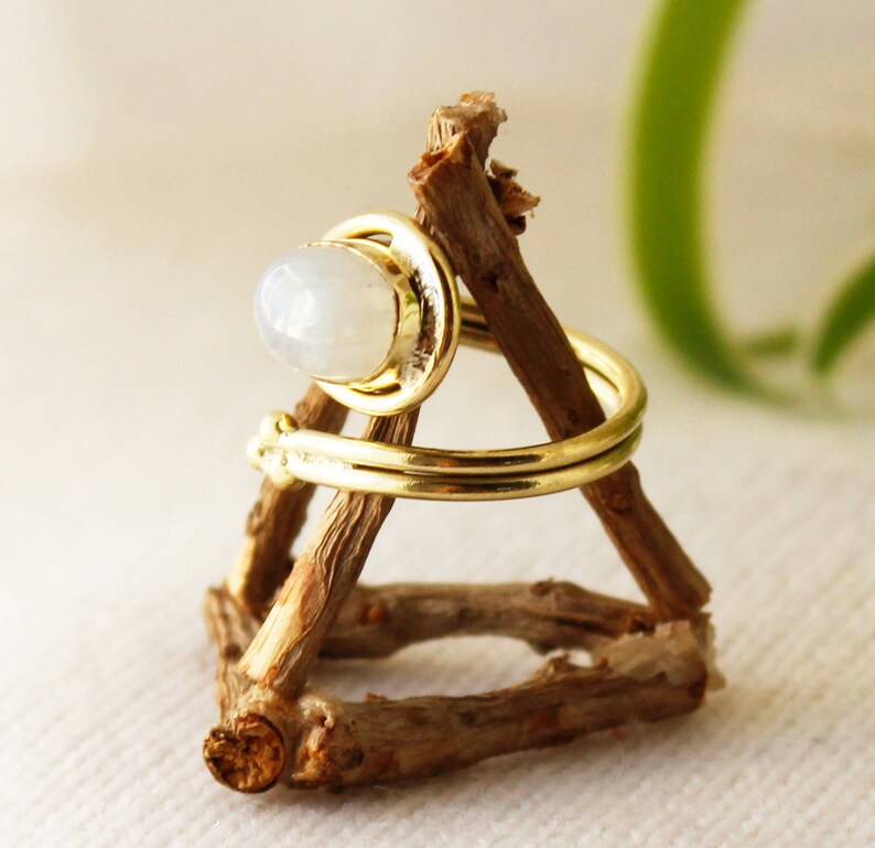 K&ouml;nnte beinhalten: Ein goldener Ring mit einem wei&szlig;en Mondstein, der in einer Zargenfassung gefasst ist. Der Ring befindet sich auf einem h&ouml;lzernen Ringst&auml;nder.