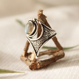 May include: A silver ring with a large, round, iridescent gemstone set in a geometric design. The ring is displayed on a wooden stand.
