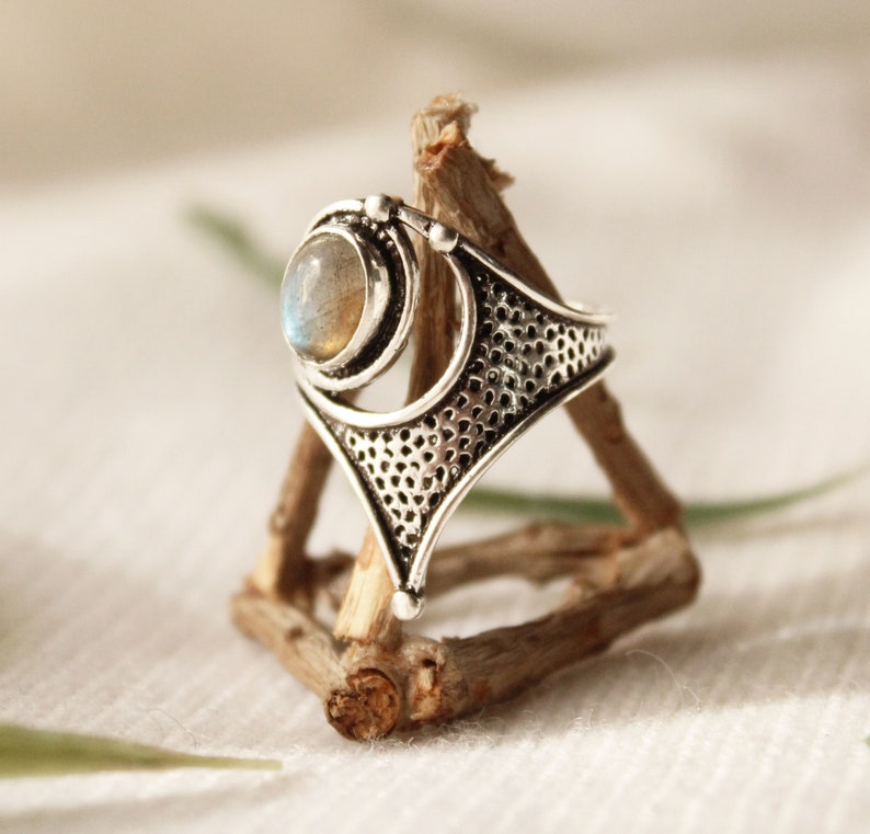 May include: A silver ring with a large, round, iridescent gemstone set in a geometric design. The ring is displayed on a wooden stand.
