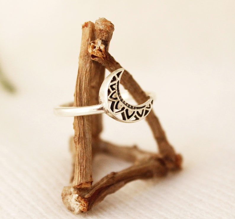 May include: A silver crescent moon ring with a detailed mandala design. The ring is displayed on a wooden stand.