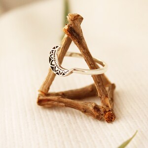 May include: A silver ring with a crescent moon design. The moon is decorated with a detailed mandala pattern. The ring is displayed on a wooden stand.