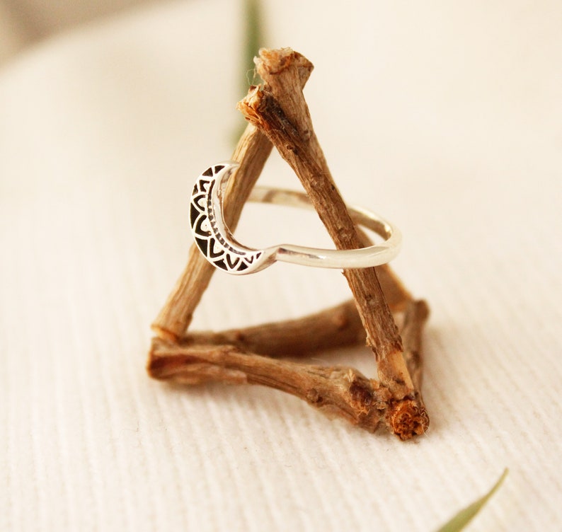 May include: A silver ring with a crescent moon design. The moon is decorated with a detailed mandala pattern. The ring is displayed on a wooden stand.