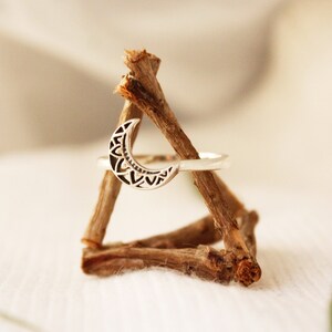 May include: A silver ring with a crescent moon design. The moon is detailed with a geometric pattern. The ring is displayed on a wooden stand.