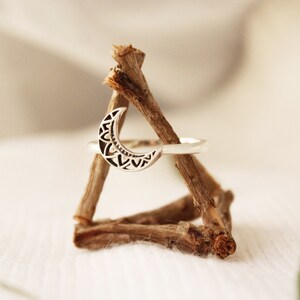 May include: A silver ring with a crescent moon design. The moon is detailed with a geometric pattern. The ring is displayed on a wooden stand.