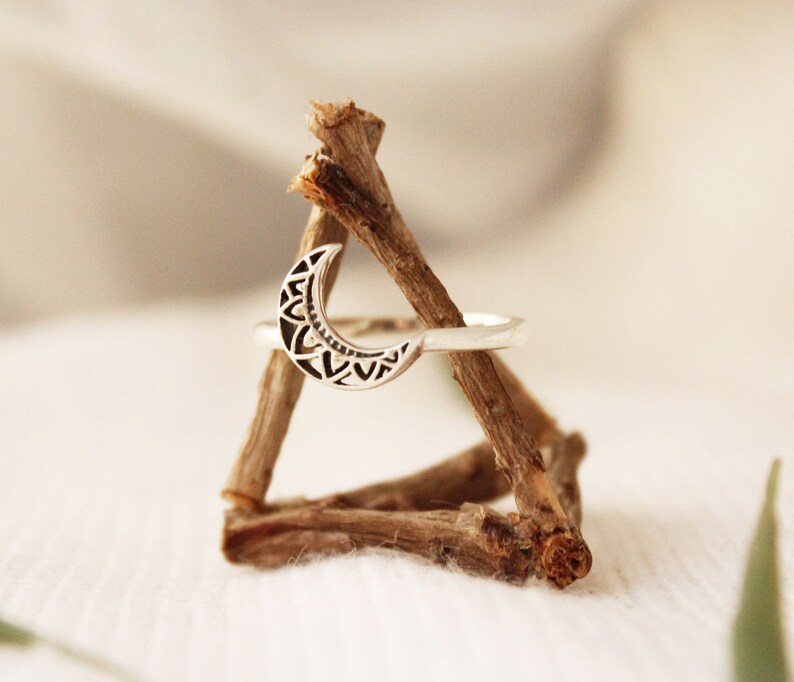 May include: A silver ring with a crescent moon design. The moon is detailed with a geometric pattern. The ring is displayed on a wooden stand.