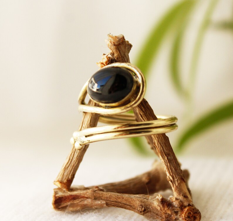 K&ouml;nnte beinhalten: Ein goldener Ring mit einem schwarzen ovalen Edelstein, der in einem drahtgewickelten Design gefasst ist. Der Ring wird auf einem Holzst&auml;nder pr&auml;sentiert.